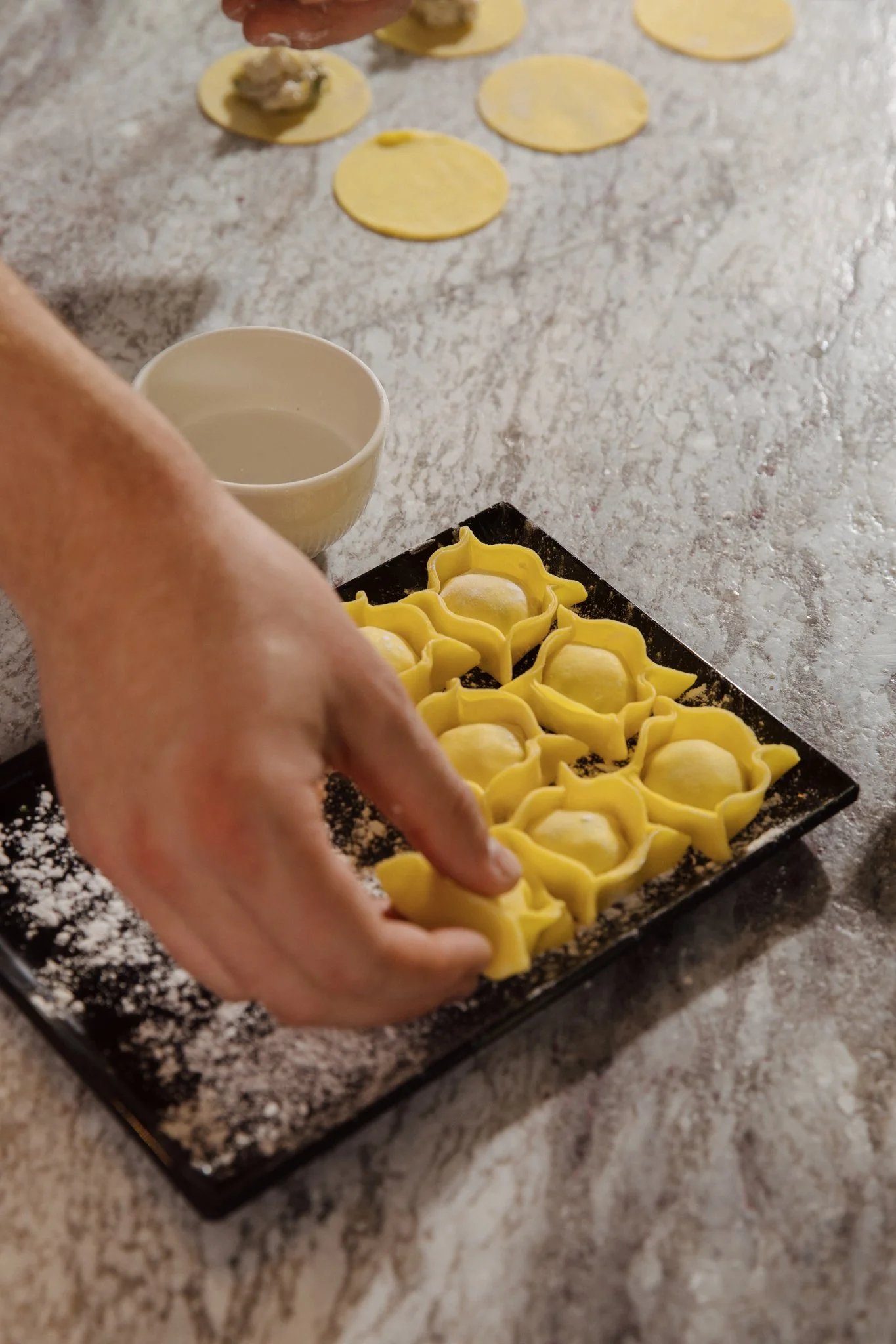 A hand laying fresh filled pasta parcels onto a dark slate tray on a marble bench